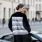 Woman wearing a navy blue jacket with a cross mink colour block back standing next to a car on a city street.
